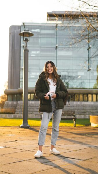 Smiling woman in a puffer jacket holding headphones, standing on a path with a modern glass building in the background.