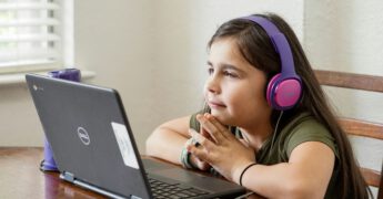 Photo of young female student with headphones on doing schoolwork on a laptop at a table.
