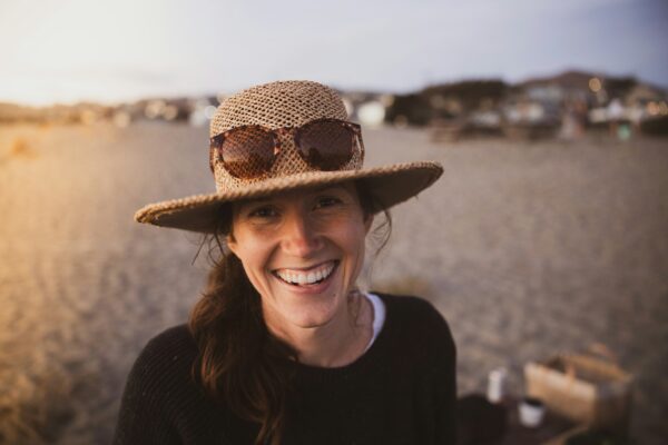 Middle-aged woman at beach with hat and sunglasses on smiling at the camera.